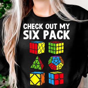 Puede incluir: Sudadera negra con el texto blanco "CHECK OUT MY SIX PACK". Debajo, seis cubos de rompecabezas coloridos en varias formas, incluyendo un cubo, una pirámide y un dodecaedro.