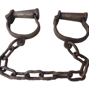 Puede incluir: Grilletes de metal antiguos con una cadena de conexión. Los grilletes tienen un diseño en forma de U con un asa y están conectados por una serie de eslabones de cadena de metal. El metal tiene una apariencia envejecida.