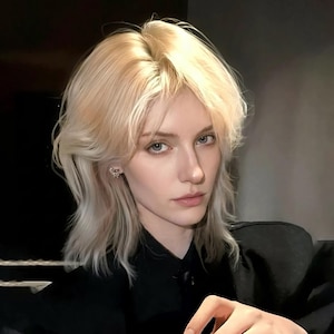 Peut inclure: Une personne aux cheveux blonds clairs et en couches, portant une chemise noire à col. La personne a la peau claire, les yeux bleus et une petite boucle d'oreille. L'arrière-plan est sombre et flou.
