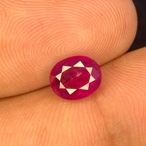 Puede incluir: Gema de rubí de corte ovalado con diseño de estrella. La gema de color rojo intenso presenta facetas triangulares blancas dispuestas en forma de estrella. El rubí se sostiene en una mano, mostrando su color y talla.