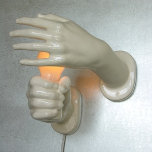 May include: A unique wall sconce featuring two porcelain hands, one open and the other clenched, holding a lit light bulb. The hands are off-white and mounted on a textured wall, with a white electrical cord.