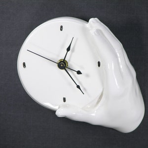 May include: White ceramic clock with black hands, shaped like a hand holding a round clock face.