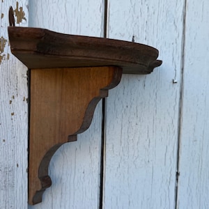Op de afbeelding: Een wandplank van hout met een halfrond bovenoppervlak en een decoratieve, uitgesneden steunbeugel eronder. Het hout heeft een medium bruine tint met zichtbare nerven en een gladde afwerking, bevestigd aan een verticale steun.