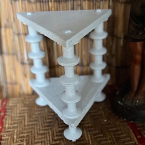 May include: A white, triangular side table with three legs. The table has a geometric design with a flat top and decorative, stacked supports. The table is painted white and sits on a woven surface.