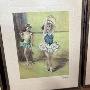 May include: Framed vintage print featuring two ballerinas in a dance studio setting. The ballerinas are wearing tutus and pointe shoes. The print has a soft, pastel color palette with a green background and a gold-toned frame.