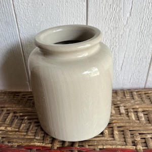Puede incluir: Un jarrón cilíndrico de cerámica con un esmalte suave y brillante de color blanco roto. El recipiente presenta un borde ancho y redondeado y un cuello corto y recto que se transforma en un cuerpo ligeramente más ancho y redondeado con base.