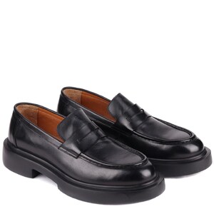 Puede incluir: Un par de mocasines de cuero negro con suela gruesa. Los zapatos tienen un dise&ntilde;o cl&aacute;sico de mocas&iacute;n con punta redonda y tac&oacute;n bajo. El interior es de color marr&oacute;n claro.