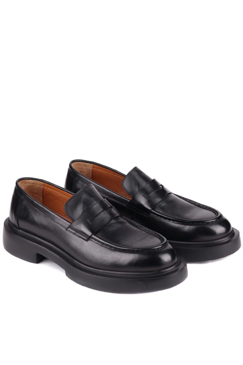 Puede incluir: Un par de mocasines de cuero negro con suela gruesa. Los zapatos tienen un dise&ntilde;o cl&aacute;sico de mocas&iacute;n con punta redonda y tac&oacute;n bajo. El interior es de color marr&oacute;n claro.