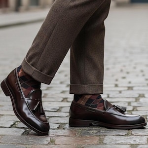 May include: A pair of polished, dark brown leather loafers with tassel detailing. The shoes are worn with brown trousers and argyle socks. The image is taken from a low angle, showcasing the shoes and the lower part of the legs.