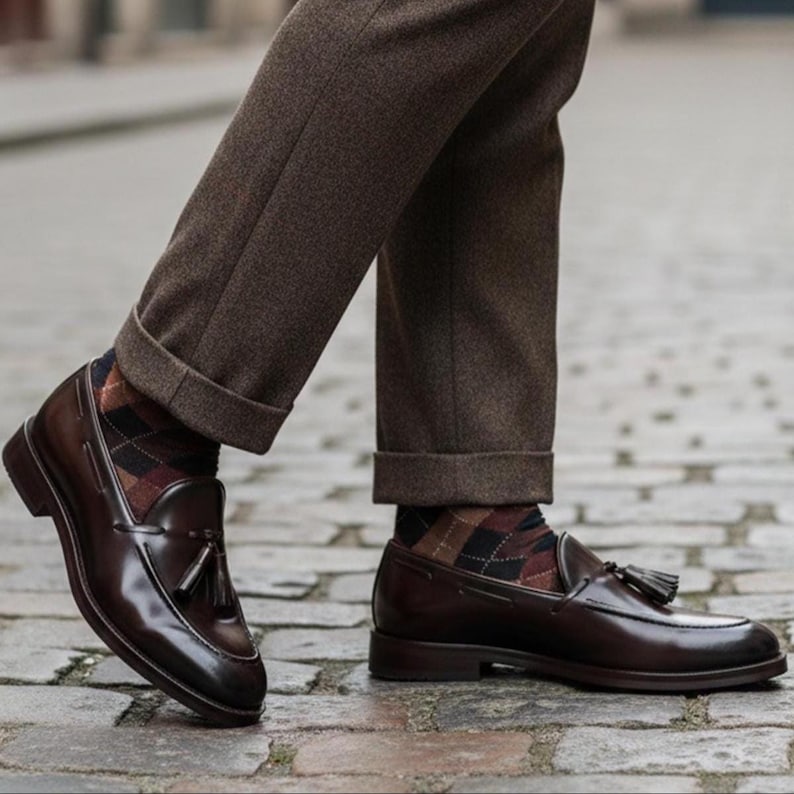 May include: A pair of polished, dark brown leather loafers with tassel detailing. The shoes are worn with brown trousers and argyle socks. The image is taken from a low angle, showcasing the shoes and the lower part of the legs.