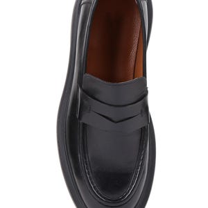 Puede incluir: Mocas&iacute;n de cuero negro con punta redondeada y suela gruesa. El zapato presenta una correa en el empeine con un dise&ntilde;o decorativo. El interior es de color marr&oacute;n claro. Un estilo de calzado cl&aacute;sico.