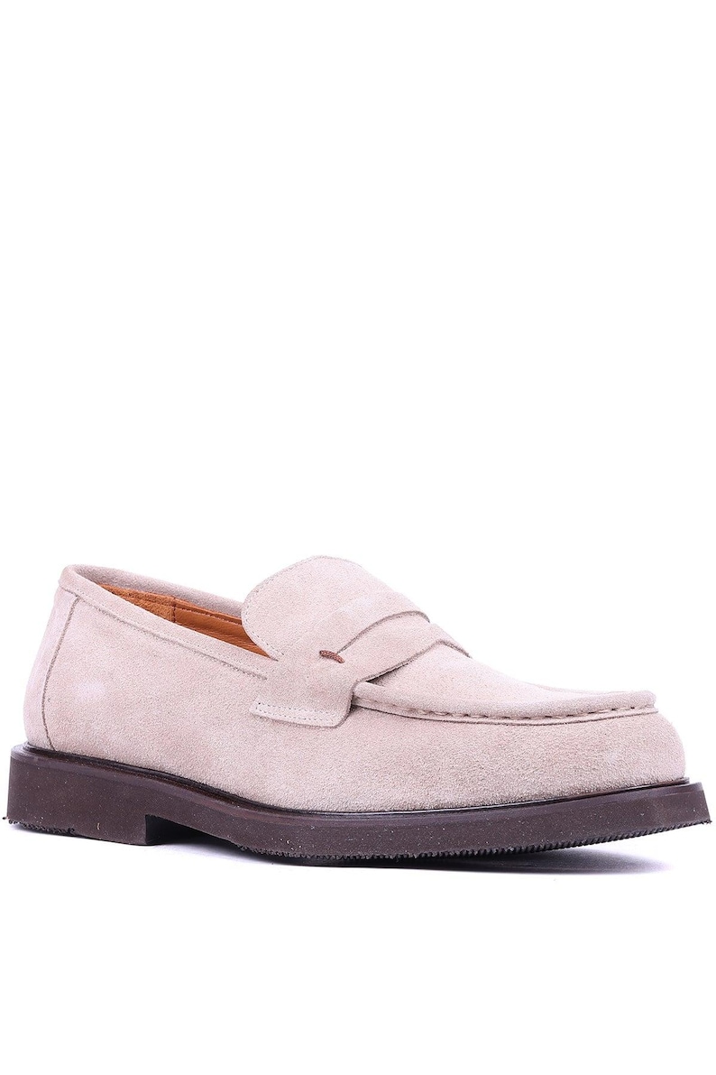 May include: A light beige suede loafer with a dark brown sole. The shoe features a classic penny loafer design with a strap across the vamp. The shoe is a neutral color and suitable for various outfits.
