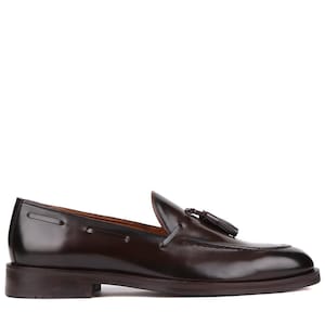 Op de afbeelding: Een enkele, donkerbruine leren loafer met een kwastje. De schoen heeft een gepolijste afwerking, een ronde neus en een lage hak. Het ontwerp is klassiek en elegant, geschikt voor formele of semi-formele gelegenheden.