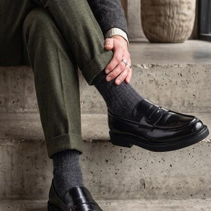 May include: Black leather loafers with a chunky sole. The shoes are worn with dark grey socks and olive green trousers. The person is wearing a tweed jacket and rings. The shoes are a classic style.