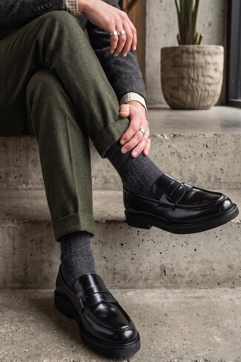 May include: Black leather loafers with a chunky sole. The shoes are worn with dark grey socks and olive green trousers. The person is wearing a tweed jacket and rings. The shoes are a classic style.