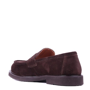 May include: Brown suede loafer with a rounded toe and a low, stacked heel. The shoe features a slip-on design with a strap detail across the vamp. The sole is a darker brown color.