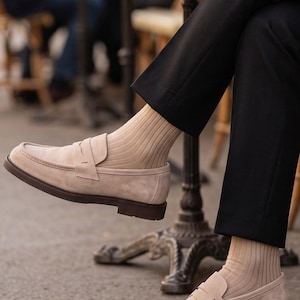 May include: Beige suede penny loafers with brown soles. The shoes are paired with tan ribbed socks and black trousers. The image is taken outdoors, suggesting a casual or semi-formal setting. The loafers have a classic, timeless design.