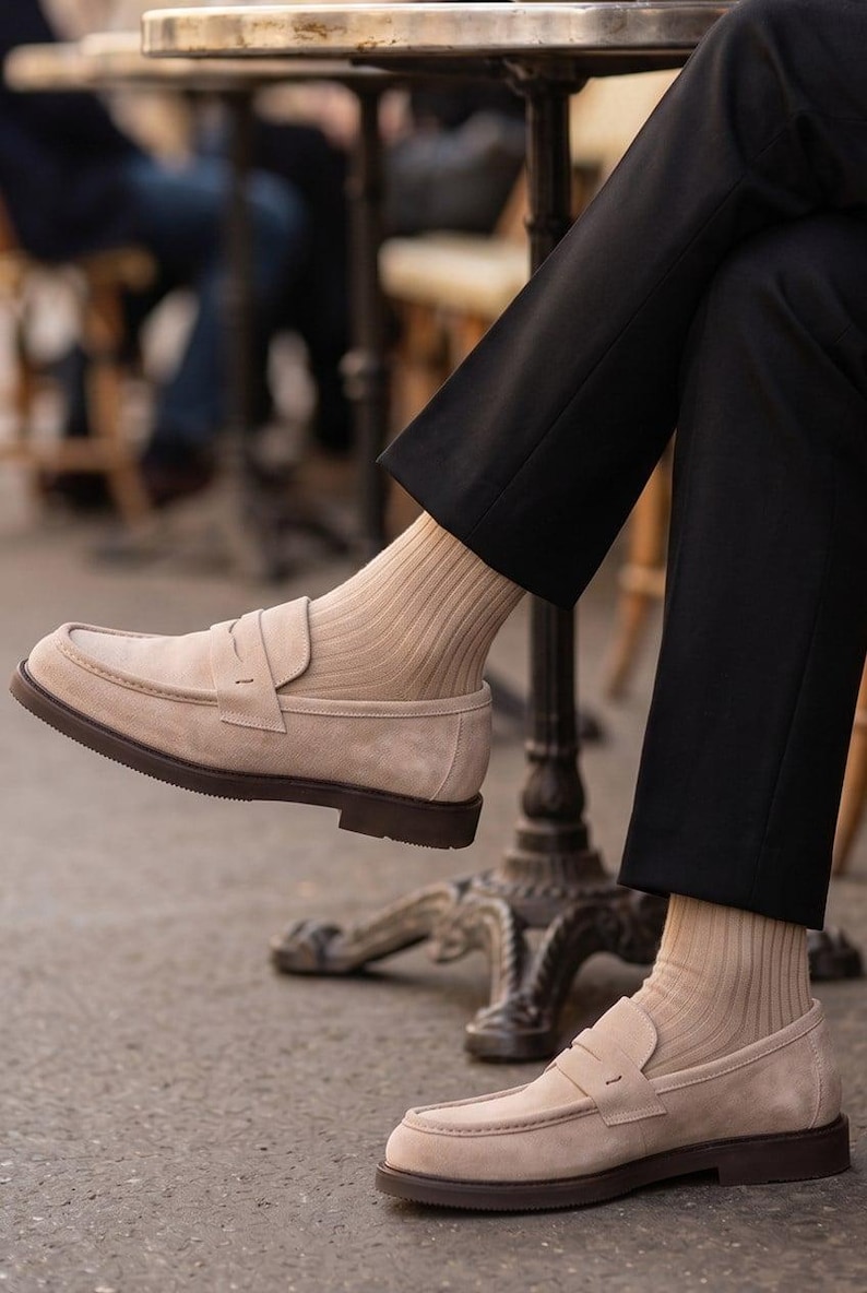 May include: Beige suede penny loafers with brown soles. The shoes are paired with tan ribbed socks and black trousers. The image is taken outdoors, suggesting a casual or semi-formal setting. The loafers have a classic, timeless design.
