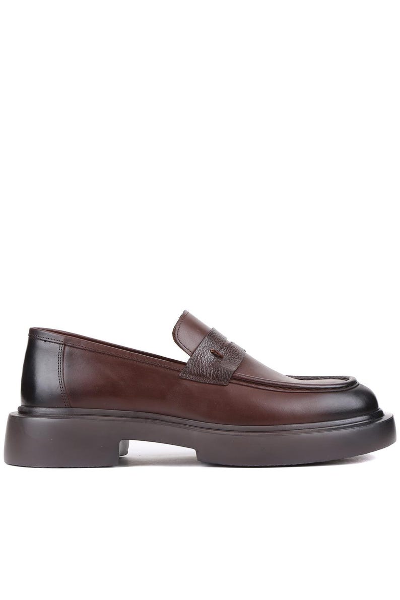 Puede incluir: Mocas&iacute;n de cuero marr&oacute;n con suela gruesa. El zapato presenta una puntera redondeada, una correa texturizada en el empeine y una suela marr&oacute;n oscuro. El tac&oacute;n mide aproximadamente 3,8 cm de alto.