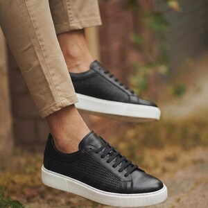 May include: Black leather sneakers with a textured weave pattern on the upper and white soles. The sneakers have black laces and are shown in an outdoor setting, suggesting casual wear.