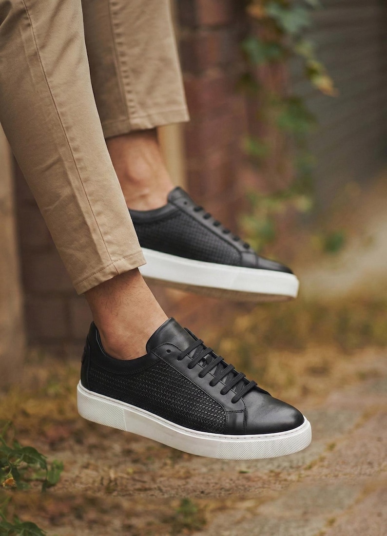 May include: Black leather sneakers with a textured weave pattern on the upper and white soles. The sneakers have black laces and are shown in an outdoor setting, suggesting casual wear.