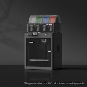 Può includere: Una stampante 3D grigio scuro con una sezione superiore trasparente che contiene quattro bobine di filamento bianche, verdi, blu e rosse. La stampante ha un display digitale e una porta. Il testo "AMS" è visibile.