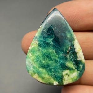 May include: A polished, teardrop-shaped gemstone featuring a vibrant blend of deep forest green, bright lime green, and creamy white marbled patterns. The surface has a smooth, reflective finish with small, dark, metallic-looking inclusions.