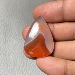 May include: A teardrop-shaped gemstone with a polished surface. The stone features layers of orange, white, and translucent hues. The gemstone is held in a hand, showcasing its size and shape. The stone's pattern creates a striking visual effect.