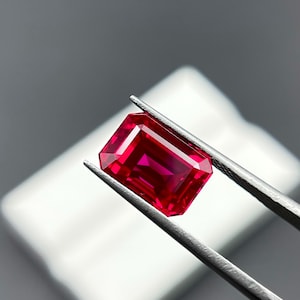 May include: A vibrant, rectangular-cut ruby gemstone held by tweezers. The gem displays a deep red hue with facets that catch the light, showcasing its clarity and brilliance. The background is a soft, neutral tone, highlighting the gemstone's color and shape.