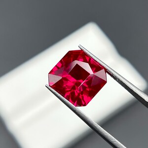 May include: A faceted, radiant-cut ruby gemstone, held by tweezers. The gemstone is a deep, saturated red color, with multiple facets reflecting light. The background is a soft, neutral gray and white.