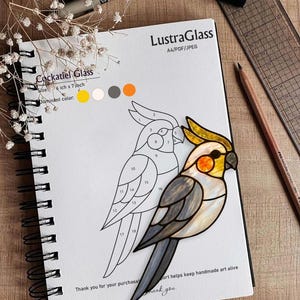 Könnte beinhalten: Ein Spiralnotizbuch mit einem Glasmalerei-Design eines Nymphensittichs. Das Design ist gelb, grau und orange. Das Notizbuch trägt auch den Text "Cockatiel Glass" und "LustraGlass". Ein Bleistift, ein Lineal und Stifte sind ebenfalls sichtbar.