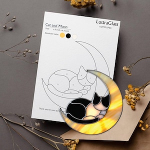 Op de afbeelding: Een glas-in-loodontwerp van een kat en maan, met een zwart-witte kat die op een gele halve maan rust. Het ontwerp is 16,5 cm bij 16,5 cm. Een papier met het ontwerp en de tekst "Cat and Moon" ligt ernaast.