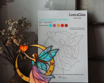 Op de afbeelding: Een glas-in-lood fee met een hart, die een hartvormig object vasthoudt, tegen een halve maan met sterren. De fee heeft blauwe en turquoise vleugels en een vloeiende jurk. De afbeelding is 20 cm bij 20 cm. Een papier met de tekst "Stained Glass Fairy with Heart" staat op de achtergrond.