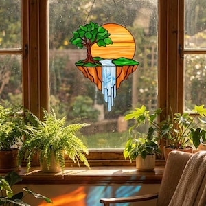Puede incluir: Vidriera que representa una escena de paisaje con un árbol, una cascada y un sol. La obra de arte está suspendida frente a una ventana, rodeada de plantas en macetas y luz natural. Los colores incluyen verde, naranja y azul.