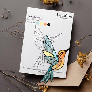 May include: A stained glass hummingbird design on a white card. The hummingbird is colored with shades of blue, yellow, and orange. The card reads "Hummingbird" and is 6 inches by 8 inches. The card is on a brown surface with dried flowers.