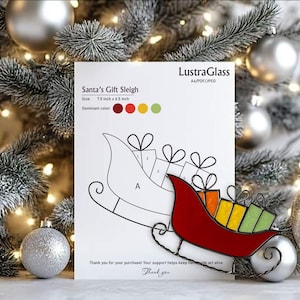 May include: A stained glass Santa's sleigh design with gifts in red, orange, yellow, and green. The design is 19.05 cm by 16.51 cm. The image is set against a Christmas tree with silver baubles.