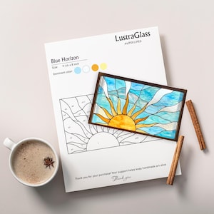 Könnte beinhalten: Ein Glasbild "Blue Horizon" mit einem Sonnendesign, 28 x 20 cm, liegt auf einem weißen Papier mit einer passenden Strichzeichnung. Das Bild enthält Zimtstangen und eine Tasse Kaffee.