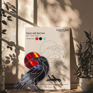 May include: A stained glass raven with a red sun, displayed on a wooden surface. The artwork is 20 cm by 20 cm. The raven is black with blue and purple accents. A drawing of the raven is in the background.