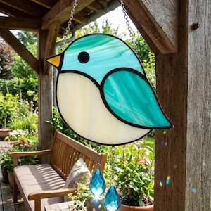 May include: A stained glass bird suncatcher with a teal and cream color scheme, hanging from a chain. The bird has a black eye and a yellow beak. Two blue crystal teardrops dangle below. The suncatcher is hanging outside.