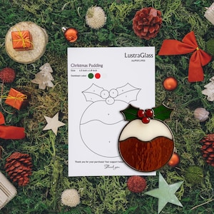 Pode incluir: Um enfeite de Natal em vitral com um pudim de Natal, com detalhes de azevinho e bagas. O enfeite mede 16,5 cm x 17,3 cm. A imagem também inclui um padrão e outras decorações de Natal.