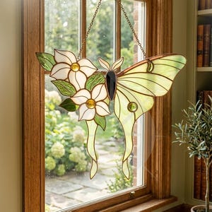 May include: Stained glass moth and flower window hanging. The moth has pale green wings with a black body and is paired with white flowers and green leaves. The artwork hangs from a chain in front of a window.