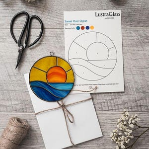 Puede incluir: Una obra de arte en vidrio que representa una puesta de sol sobre el océano, de 16,5 cm por 16,5 cm. La obra presenta un sol amarillo y naranja, olas azules y una guía de patrones. El texto "Sunset Over Ocean" es visible.