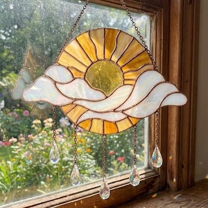 Puede incluir: Atrapasoles de vidrio manchado con un sol amarillo con rayos y detalles de nubes blancas. Colgantes de cristal transparente en forma de lágrima cuelgan debajo. El atrapasoles está suspendido por una cadena.
