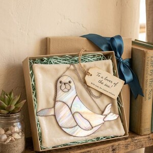 Peut inclure: Un pendentif en vitrail représentant un phoque, avec une chaîne argentée, présenté dans un coffret cadeau avec une étiquette indiquant "To a lover of the sea!". Le phoque est blanc, avec des reflets irisés.
