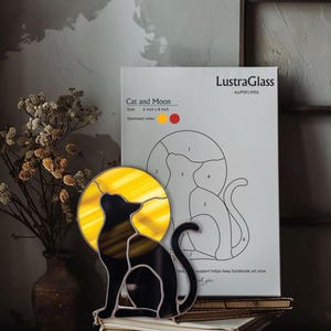 May include: A stained glass cat silhouette with a yellow moon, 6 inches by 8 inches, sits atop a stack of books. The design is black and yellow. A pattern for the cat and moon is visible in the background.