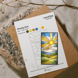 May include: A stained glass panel with a sunrise design, measuring 5 x 10 inches, is displayed on a white card. The card includes the text "Morning Glow" and "LustraGlass". A brown envelope and a stone are in the background.