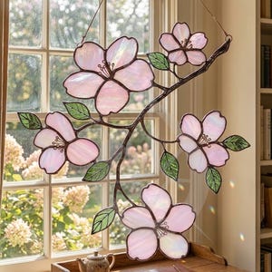 May include: A stained glass floral artwork showing five pink blossoms and green leaves on a brown branch. The artwork hangs in front of a window, with the text "LustraGlass" in the upper left corner. A decorative window hanging.