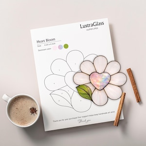 May include: A stained glass flower with a heart-shaped center, measuring 6 inches by 6 inches, rests on a paper with a flower outline. The paper includes the text "Heart Bloom" and "LustraGlass". A cup of coffee and cinnamon sticks are also present.