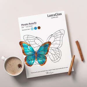 May include: A detailed illustration of a Morpho Butterfly, 9 inches by 8 inches, with a vibrant teal and brown color scheme. The image is on a white paper with the text "LustraGlass" and "Morpho Butterfly". A cup of coffee and cinnamon sticks are also present.