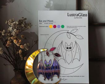 Puede incluir: Decoración de vidriera de murciélago y luna. El murciélago tiene alas moradas y ojos verdes, colgando de una luna creciente dorada. Tamaño: 20 cm x 20 cm. Un papel con el texto "Bat and Moon" y "LustraGlass" está detrás de la decoración.